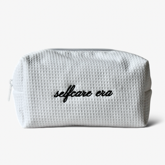 Wit make-up tasje van zachte wafelstof met geborduurde tekst ‘selfcare era’.